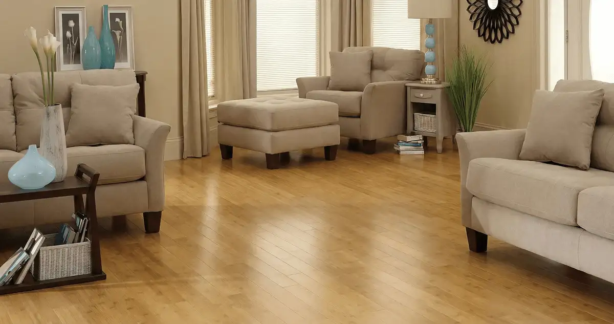 A living room with professionally installed bamboo flooring, beige sofas, a matching ottoman, a side table with a blue lamp, vases, framed pictures, and large windows with blinds and curtains in Eugene, OR
